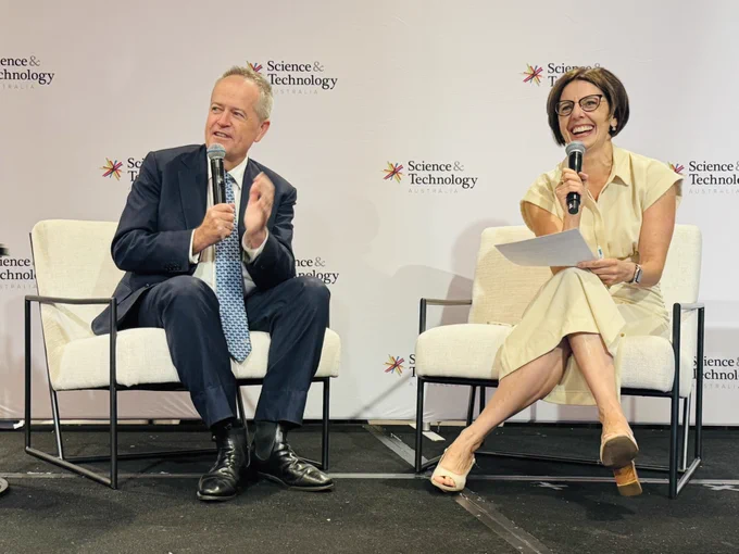 Two people sit on stage on chairs, while holding microphones, one speaking and one smiling. The logo of Science and Technology Australia is behind them. 