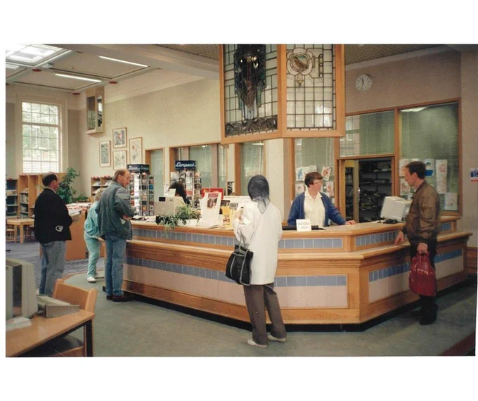 A photograph of a library counter, with people working behind it, and people standing waiting to be served.