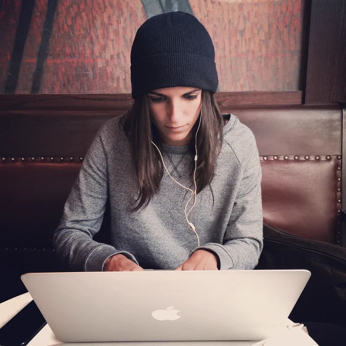 A woman wearing a navy beanie hat and grey jumper working on asilver laptop.