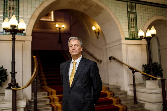 Photo of Richard Atkinson. White man with grey hair wearing a black striped suit, yellow tie and white shirt standing by some stairs in the Law Society. 