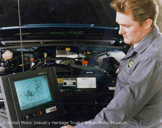 A colour photograph of a male technician standing in front of an open car bonnet and looking down at a Testbook computer next to him. The computer's screen displays a schematic of an engine part. 