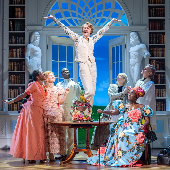 The cast of The Importance of Being Earnest surround a man wearing all white who stands on a table with his arms in the air.