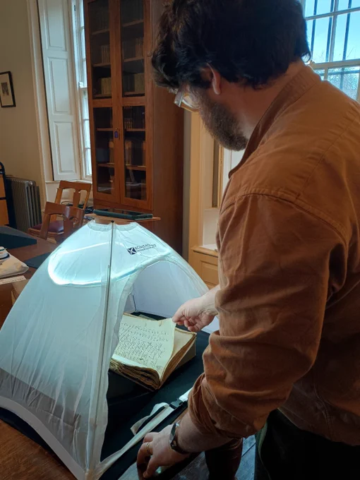 Photograph of a man standing and turning the page of a manuscript that is resting on supports in a 'scantent' with inbuilt lights
