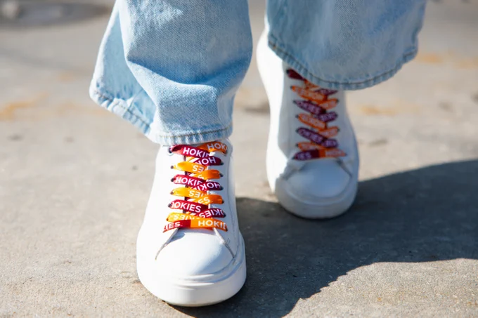 A close-up photo of a pair of white sneakers with custom shoelaces in alternating maroon and orange colors, featuring the word "HOKIES" printed repeatedly in white text. The wearer is standing on a concrete surface, with sunlight casting a shadow behind them. The shoelaces are arranged in a crisscross pattern, creating a vibrant and eye-catching design.