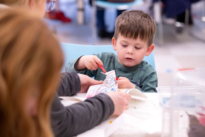A child with short brown hair and a green sweatshirt cutting a piece of paper with scissors.