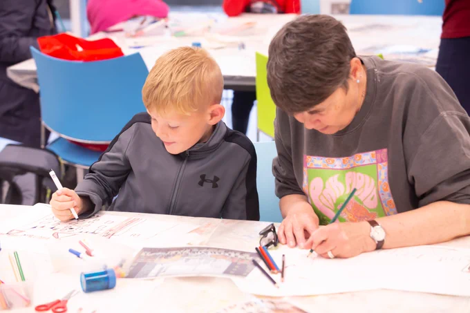 An adult and child sat at a table with pens, pencils and paper. The child has short blonde hair and a grey and black zip up top. The adult has short dark hair and is wearing a grey sweatshirt with a colourful logo.
