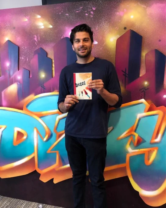 Playwright Mohamed-Zain Dada holds a copy of the Dizzy playtext, stood in front of a large, brightly coloured graffiti-style graphic displaying the words 'Dizzy'. 