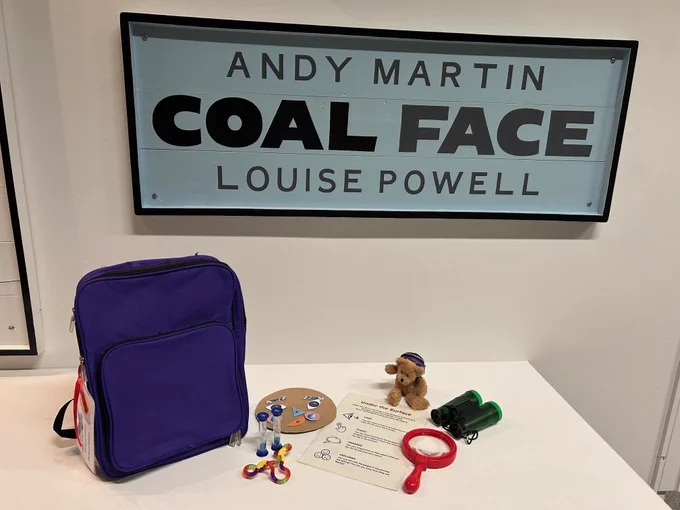 A blue and black painted sign which reads 'Andy Martin, Coal Face, Louise Powell'. On the white table below the sign is a purple backpack. Besides the backpack is a selection of colourful objects, including a magnifying glass, binoculars and a small soft toy bear.