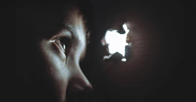 A person looks through a hole ripped into a wall at a bright light.