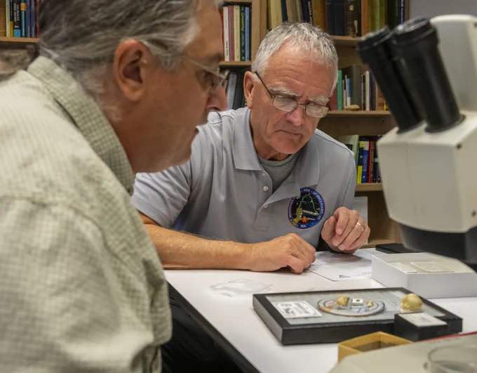 two men are at a desk with a microscope looking at a tray of some kind holding small objects and papers