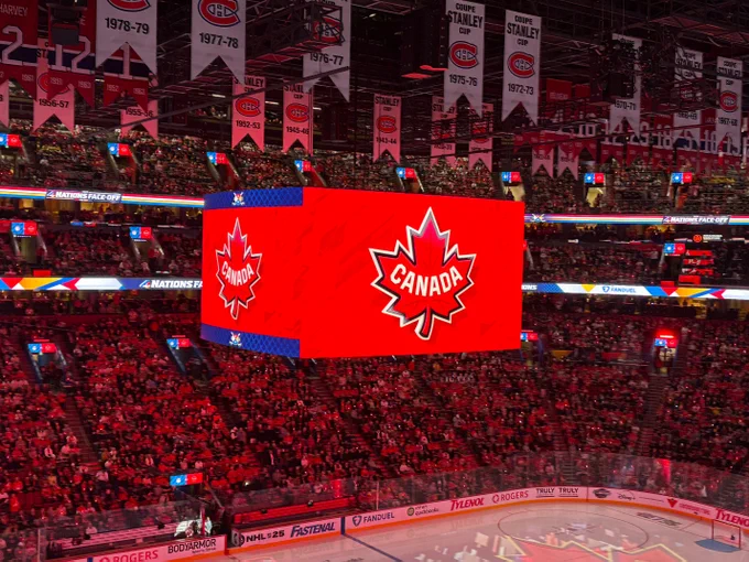A hockey arena is lit up in red and filled with spectators. The Team Canada 4 Nations Face-Off crest is displayed on the jumbotron.