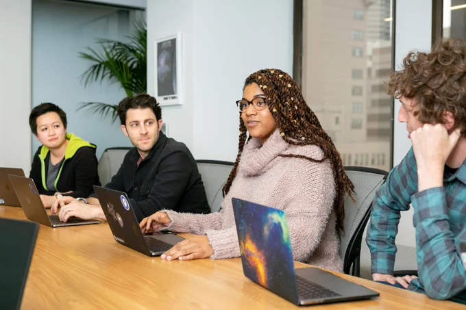 a photograph of young people sitting at a conference table with laptops 