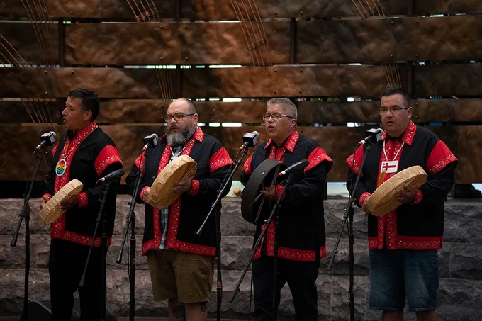 Four men in matching red and black shirts perform on stage, singing and beating circular hand drums.