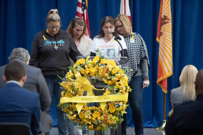 people speaking behind a podium and a wreath of yellow flowers