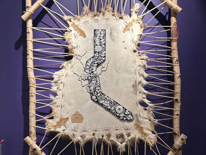 A picture of the "Bead the Tract" beaded display. In front of a purple background wall a cleaned animal skin is stretched out on a wooden frame made of debarked branches. Dozens of thing strips of leather attach the skin to the wood. In the middle of the skin, purple and white beads are arranged in the shape of the Haldimand Tract (shaped like a long tube sock kinked out to the left then right). Image: The City of Waterloo Museum.