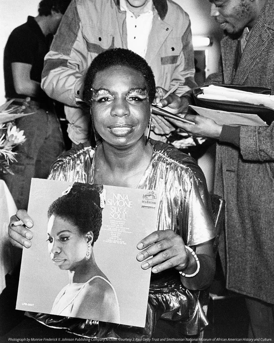 A black-and-white photograph of Nina Simone holding a vinyl record cover of 'Nina Simone Sings the Blues' while seated indoors, wearing a shimmering silver jacket and smiling at the camera. Circa, 1967.