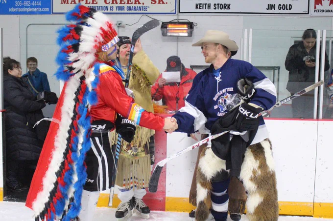 The photo shows two individuals shaking hands on an ice rink. One person is dressed in traditional Indigenous regalia, including a colorful feathered headdress and red outfit, while the other wears a hockey uniform with a blue jersey and furry chaps, holding a hockey stick. Spectators and signs, including one for "Maple Creek, SK," are visible in the background, indicating a cultural or community hockey event.