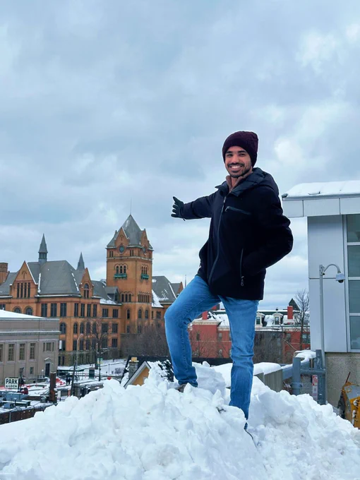 Individual posing on a snowy mound with a backdrop of the historic building of Wayne State University's Old Main.