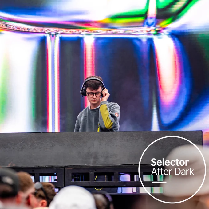 DJ Reger performing on stage at AVA Festival, standing behind the decks with one hand on his headphones. The crowd is in front of him.