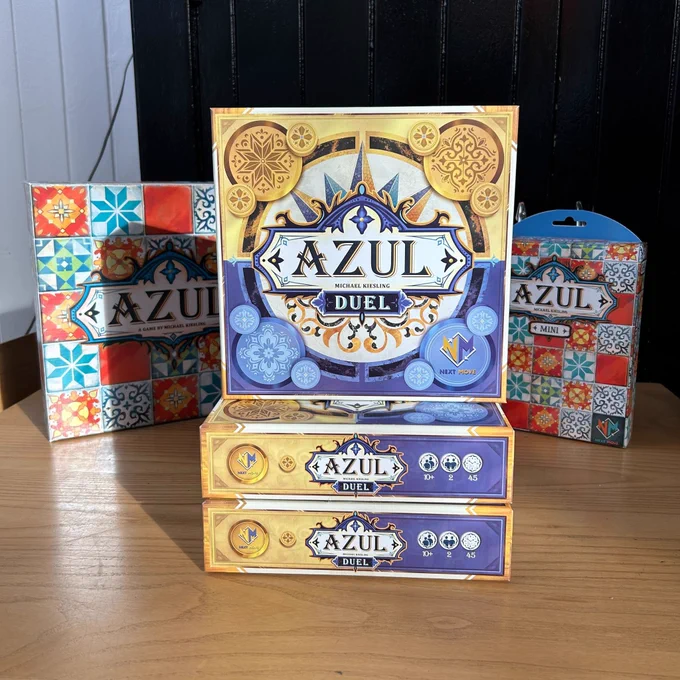 Three board games in the Azul series on a table in the board game shop Ancient Robot Games. In the middle are copies of the new Azul Duel with a yellow and purple cover. Behind the Azul Duel there are copies of the original Azul and Azul Mini.