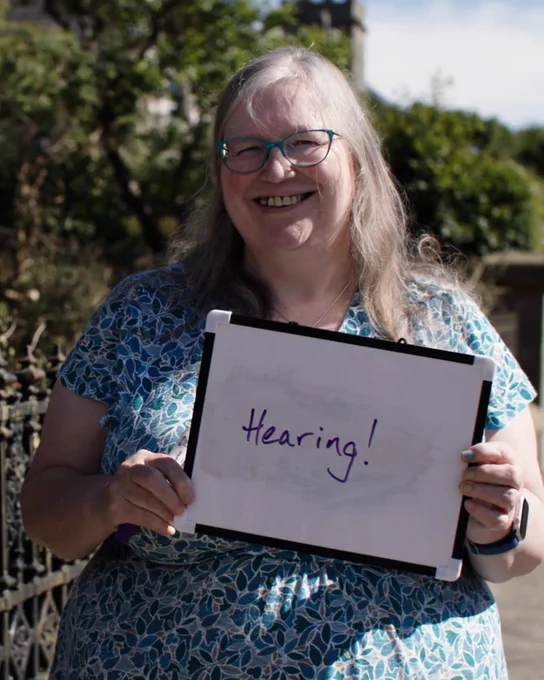 A woman standing, smiling and holding a board reading "hearing!".