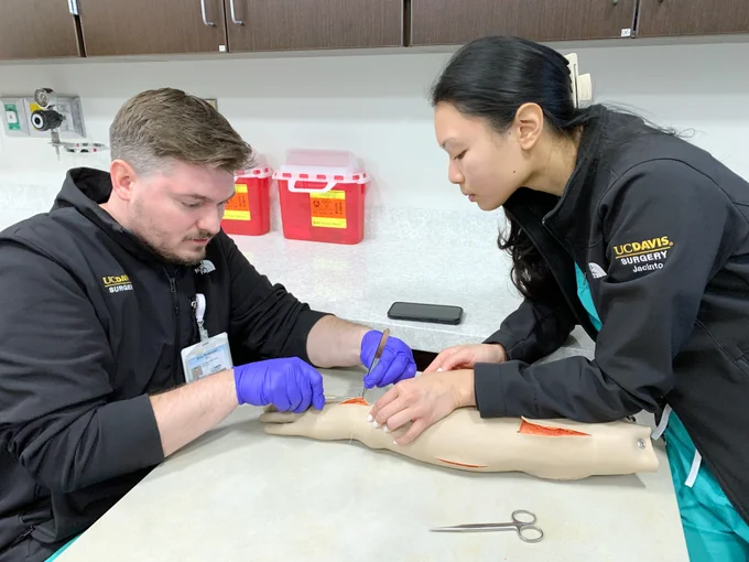 Two surgery residents suture a task-trainer simulated manikin arm.