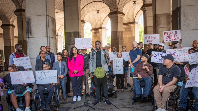 The Public Advocate speaks at a rally outside of the Dinkins building, as Council Members and advocate listen. Advocates, standing and seated in wheelchairs, hold signs with messages like "People Need CDPAP" and "Family Care is a job"