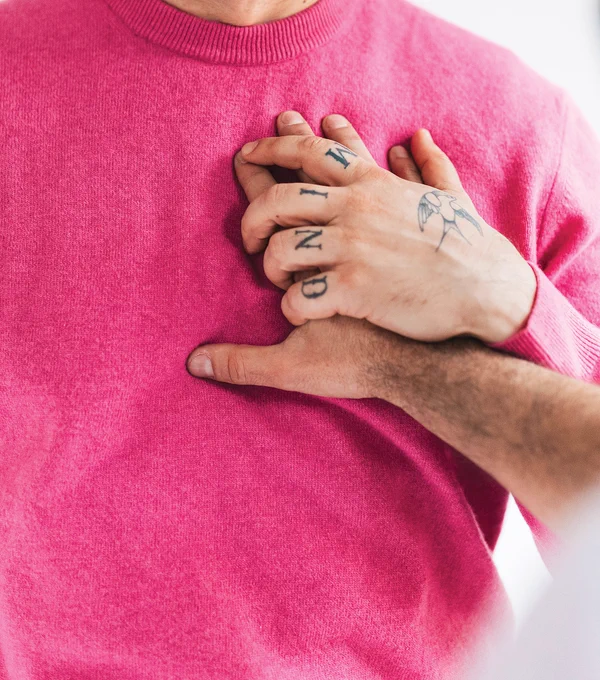 Close-up photo of a person in a pink sweater with a tattooed hand holding another person's palm to their heart