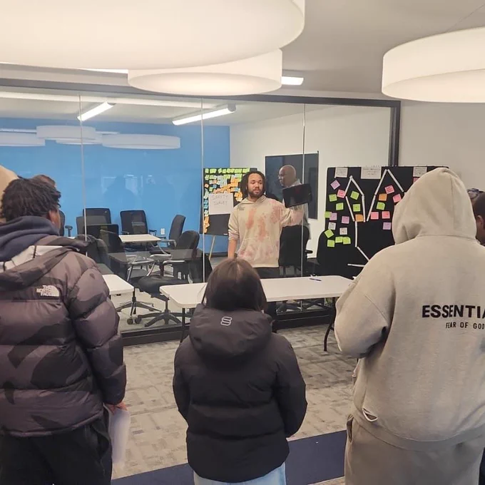 ID 1: A group of people listening to a speaker through a glass wall in a modern room with sticky note-covered boards.