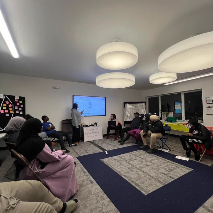 ID 2: A group of people seated in a circular formation during a meeting or workshop in a modern room with a screen, light fixtures, and a whiteboard.