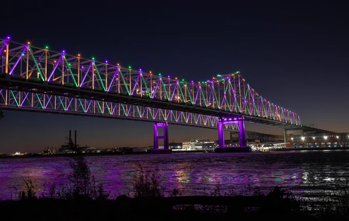 A night view of the illuminated Crescent City Connection bridge over the Mississippi River in New Orleans. The lights are purple, green, and yellow.