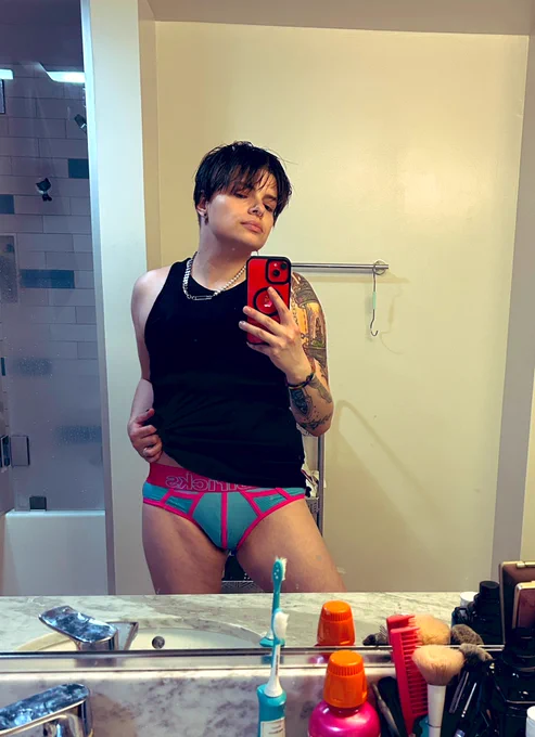 Micki lifting their tank top to better show off neon pink and blue briefs