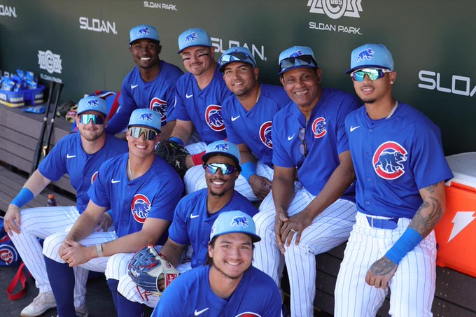 Sammy Sosa poses in the Cubs dugout with various Cubs players and prospects. They wear blue Spring Training jerseys and caps with white baseball pants.