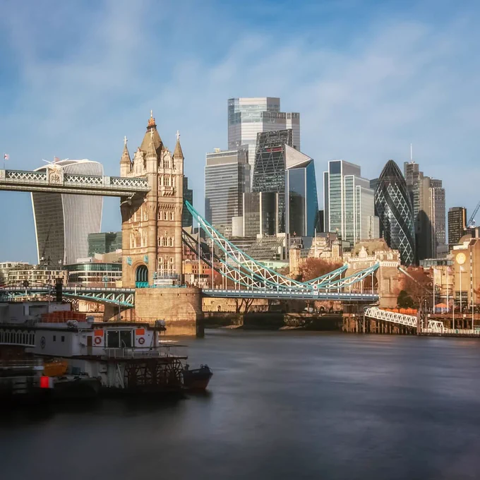The north side of Tower Bridge is pictured in the sun. The Cities skyscrapers are in the background against a blue sky with fluffy clouds. 