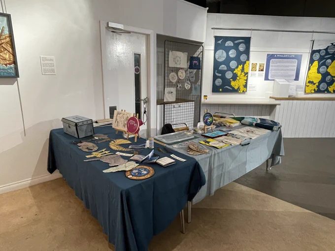 Two cloth covered tables covered with embroidered textiles and handmade books stands in a gallery space with hung textile panels behind.