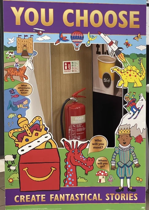 A  floor standing graphic panel acting as a frame with a cut out centre has cartoon images of a dragon,  a dinosaur, downhill skier, mermaid, castle and other fantasy imagery .  It has been propped in a corner so it frames a fire extinguisher and an image of a cup of coffee.