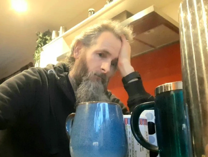 Photo of Darren in kitchen resting his head on his hand looking exhausted. Cups in foreground.