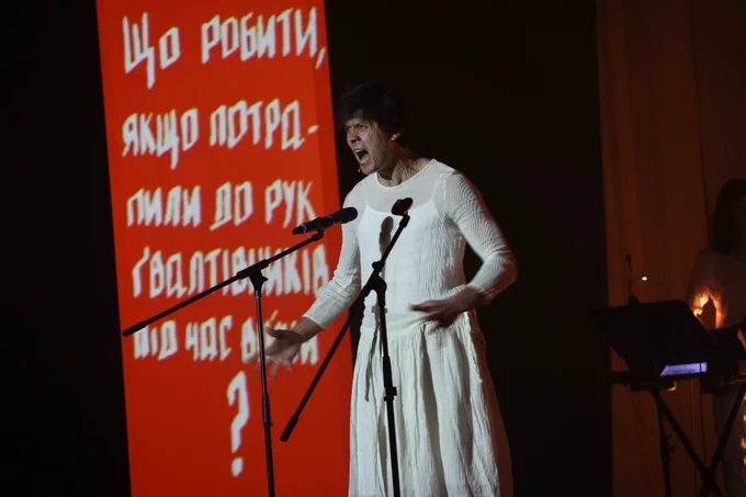 Szenenfoto mit einer Person in einem weißen Kleid, die in ein Standmikro singt - dahinter eine Projektion mit weißer Schrift auf rot