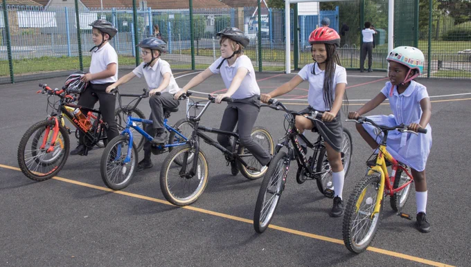 A group of five children in school uniforms, wearing helmets, sit on their bikes in a playground during a Bikeability Level 1 training session. They appear focused and ready to practice essential cycling skills in a safe, enclosed space.