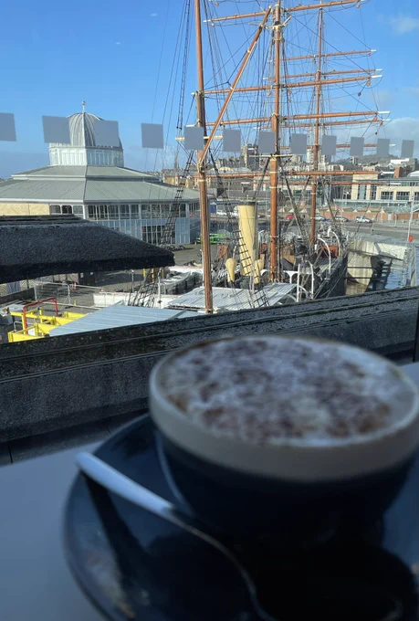 In the foreground is an out of focus cappuccino in shadow. Through the window is a view looking down on the RRS Discovery bathed in sunshine.