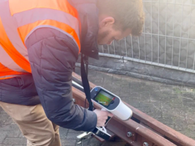 A person wearing a high-visibility orange vest and a padded jacket inspects a rusted rail using a handheld scanning device. The device's screen displays data, suggesting a structural or material analysis. 