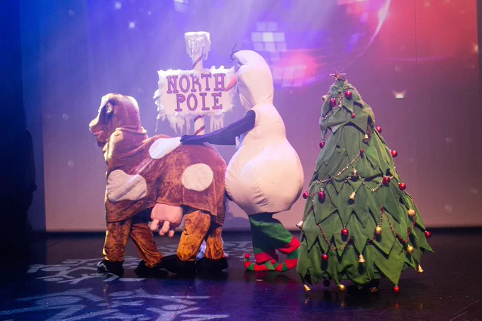 A pantomime cow, snowman and Christmas tree dancing in a line