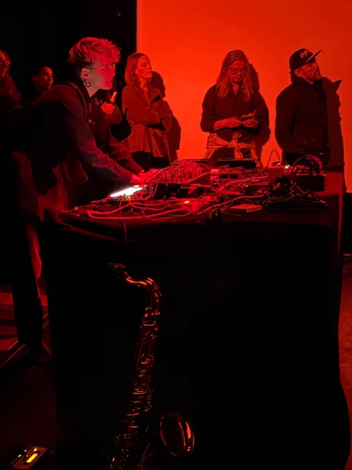 Sonia Killmann performs on a modular synth. They have short blonde hair and are wearing a black boiler suit. Audience members surround them. The lighting is red. 