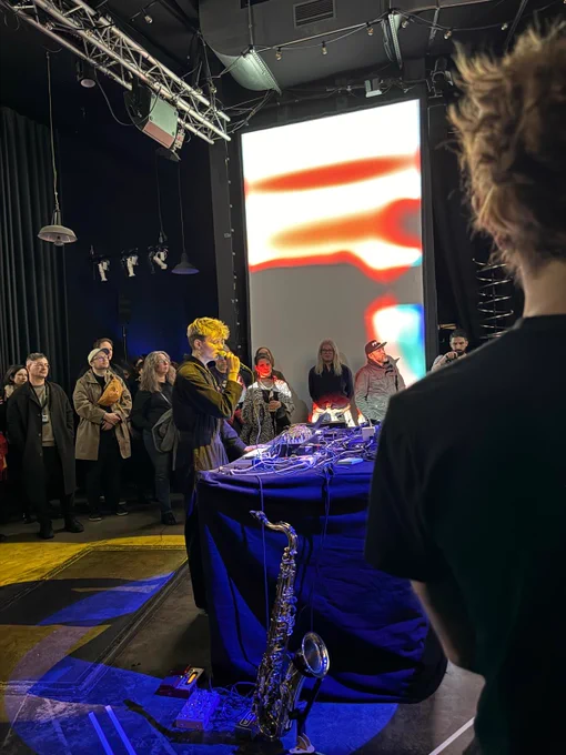 Sonia Killmann sings into a microphone. They have short blonde hair and are wearing a black boiler suit. Audience members surround them