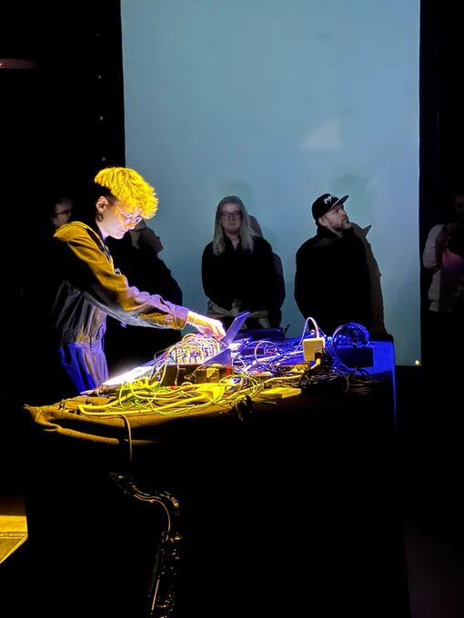 Sonia Killmann performs on a modular synth. They have short blonde hair and are wearing a black boiler suit. Audience members surround them