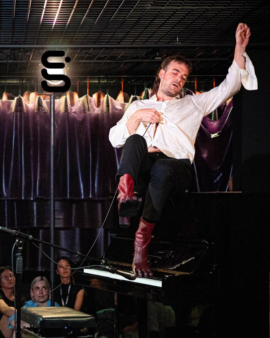 A performer sits on a piano, wearing red leather shoes.