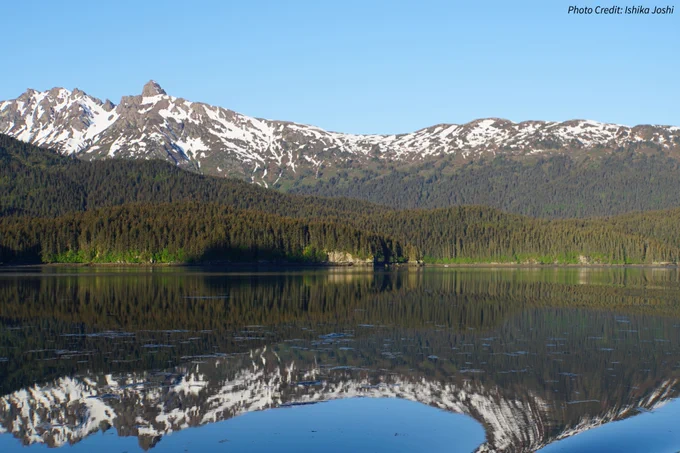 Glassy water reflects tree and snow covered mountains in the distance.