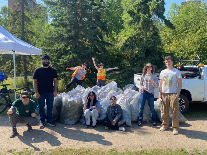 Six people sit or stand in front of a pile of clear garbage bags full of plant material. Two people in high vis vest stand behind the pile, looking excited. They are in a park and it is very sunny. 