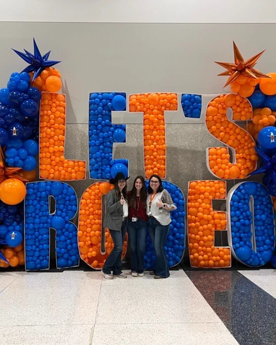 Three people standing in front of a large decorative sign that reads "LET'S RODEO" adorned with blue and orange balloons.