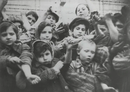 A black and white historical photo showing a group of children in stripe concentration camp uniforms reaching out towards the camera, displaying their forearms their tattooed numbers. After liberation of Auschwitz. 